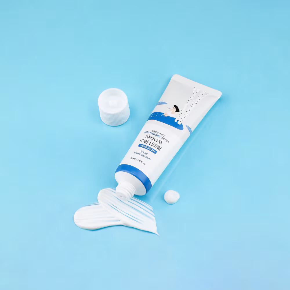 A Round Lab Birch Juice Moisturizing Sunscreen tube with blue accents lies on a light blue background, with white cream squeezed out in a smear and a small dollop beside it. The cap is placed separately nearby.
