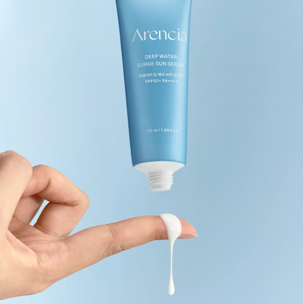 A hand is shown below a light blue tube of Arencia Deep Water Surge Sun Serum, with a single drop of white serum dripping from the tube against a soft light blue background.
