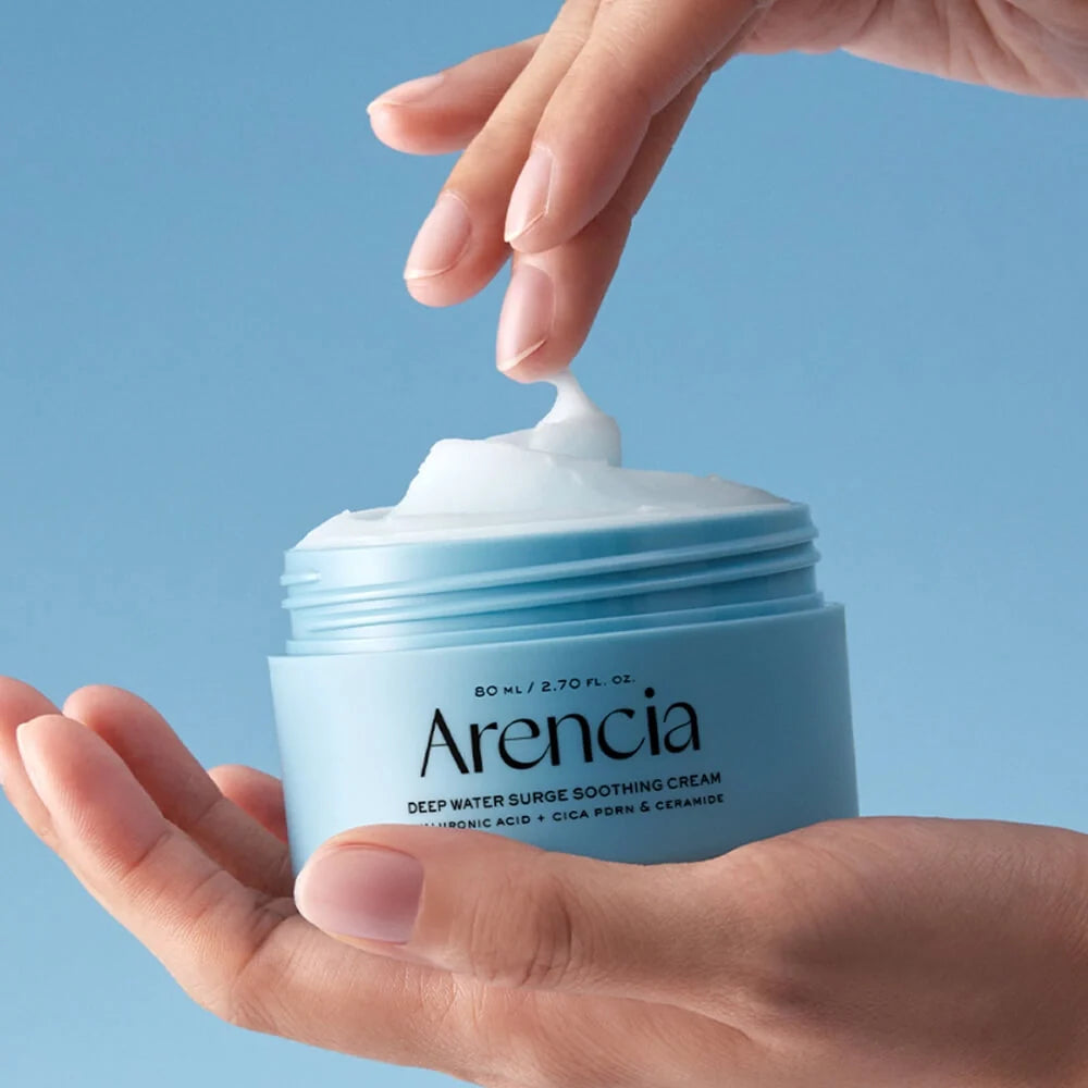 A person holds an open jar of Arencia Deep Water Surge Soothing Cream, with a dollop of white cream being spread onto their finger against a solid light blue background.
