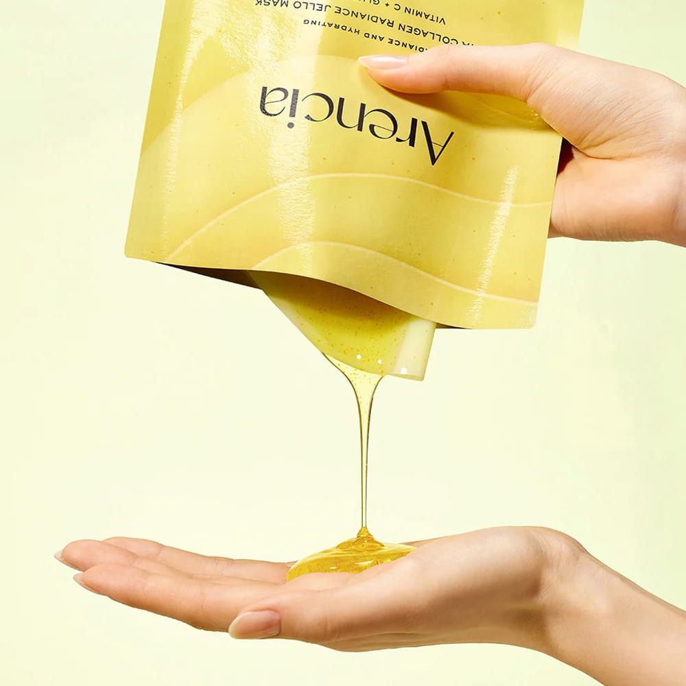 A hand holds a yellow pouch of Arencia Vita Collagen Radiance Jello Mask, pouring a thick golden-yellow gel into the other hand against a pale yellow background.