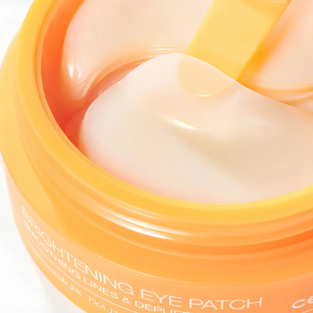 Close-up of a Celimax TXA Brightening Eye Patch containing smooth, pale cream eye patches, with a spatula lifting one patch. Text on the jar reads 'BRIGHTENING EYE PATCH' and 'SMOOTHING LINES & DEPILEATED'.