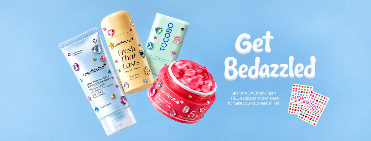 Four skincare and beauty products floating on a light blue background with the text 'Get Bedazzled' and a small sticker graphic. Products include a white tube, a yellow bottle, a mint green tube, and a pink jar of glitter or powder.