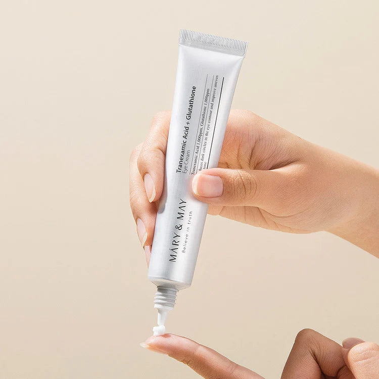 Hand holding a tube of Mary&May Tranexamic Acid + Glutathione Eye Cream with a beige background