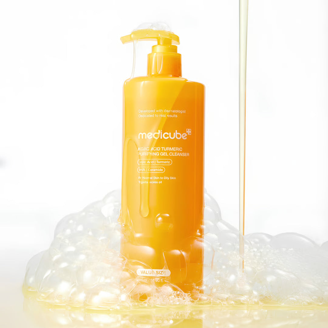 A bottle of medicube Acid Turmeric Cleansing Gel stands on a reflective white surface surrounded by foam, with a stream of yellow gel running vertically on the right side.