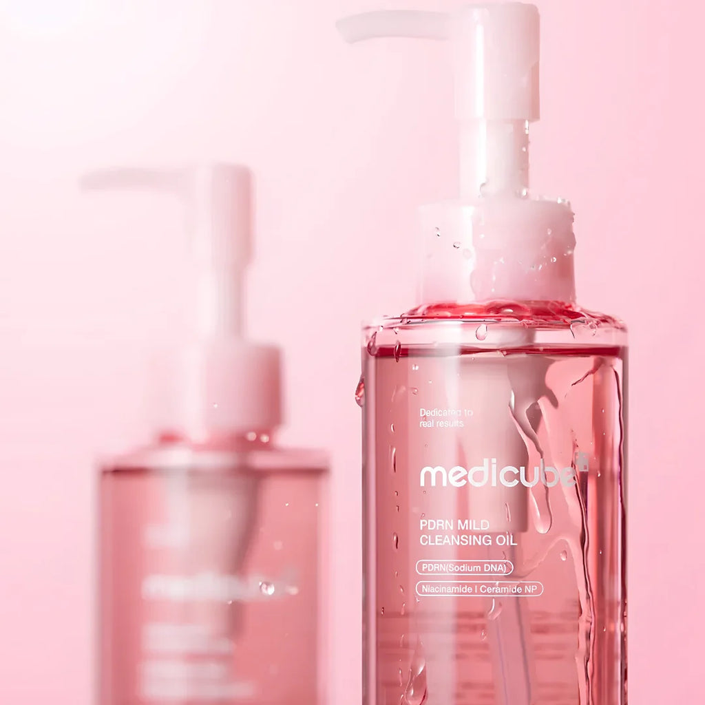 A close-up of a Meducube PDHN Milk Cleansing Oil bottle with a pump dispenser, filled with pink liquid and shown with water droplets. A second, out-of-focus bottle is visible in the background against a soft pink gradient backdrop.