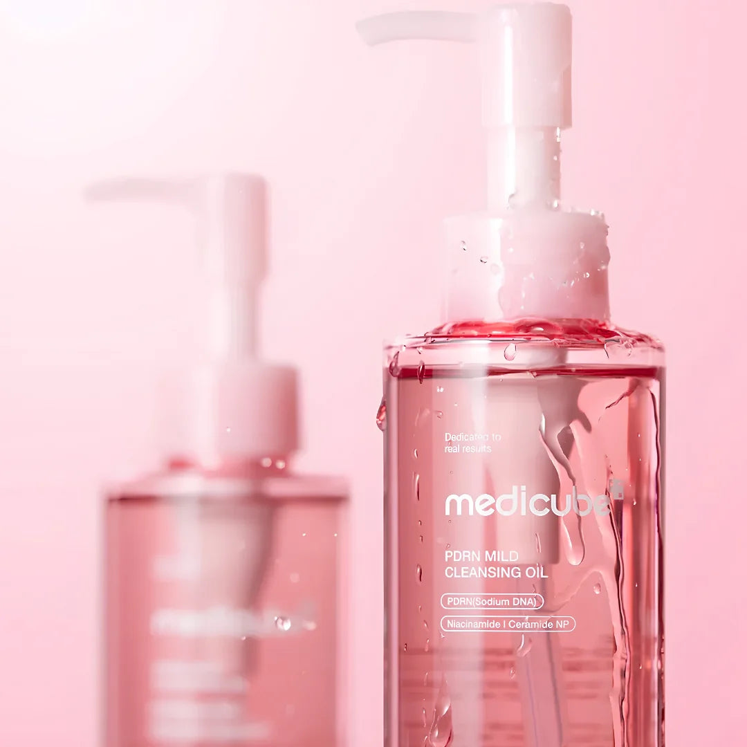 A close-up of a Meducube PDHN Milk Cleansing Oil bottle with a pump dispenser, filled with pink liquid and shown with water droplets. A second, out-of-focus bottle is visible in the background against a soft pink gradient backdrop.
