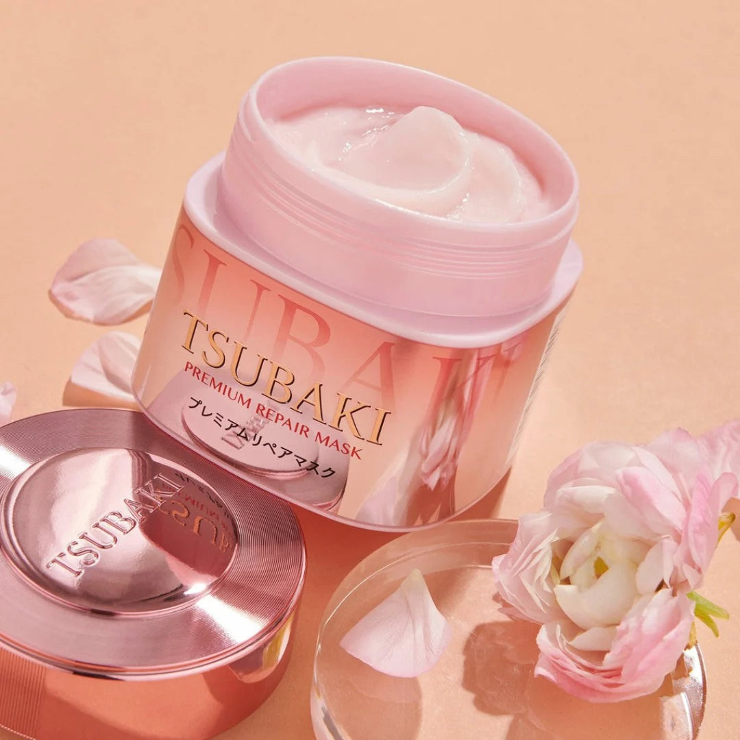 A jar of Tsubaki Premium Repair Hair Mask Pink Camellia with its lid beside it, styled with pink flower petals on a peach background.