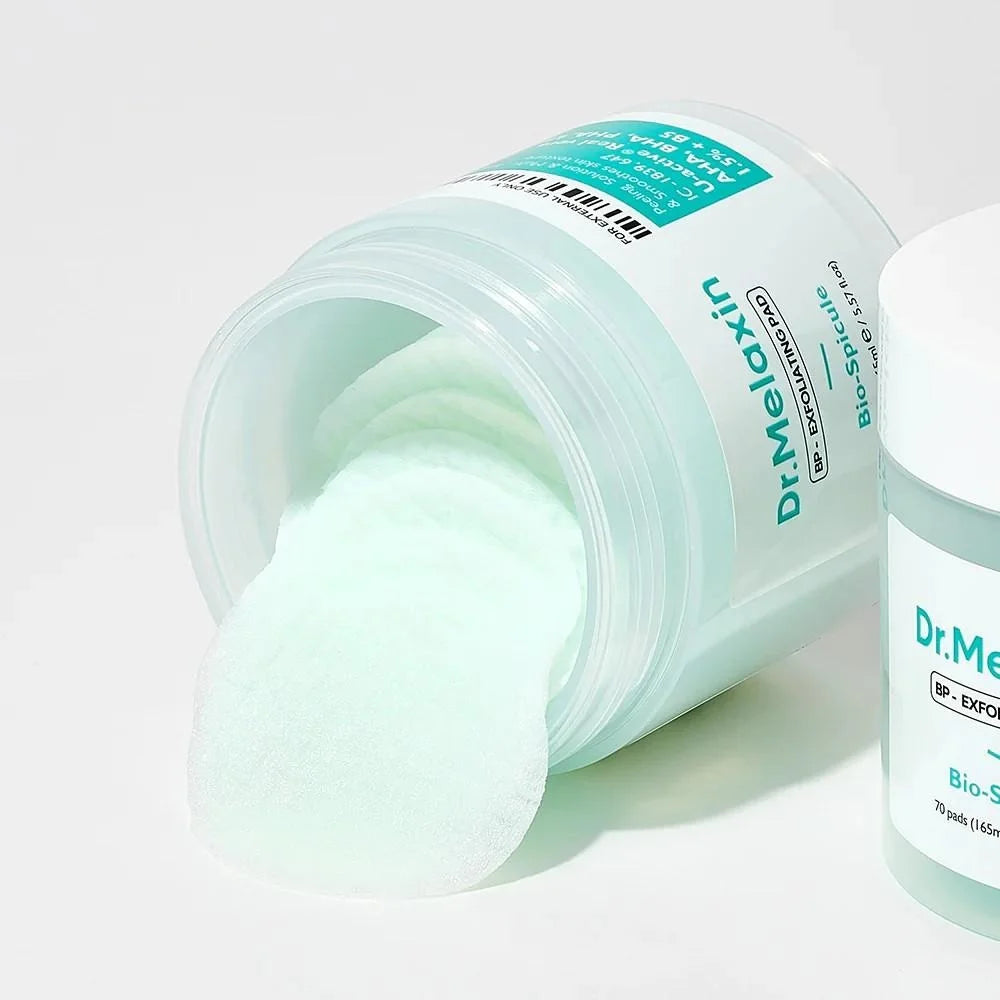 A clear plastic jar of Dr.Melaxin Bio-Spicule Exfoliating Pad 70 Pads tipped over on a white surface, with pale mint-green cotton pads spilling out.