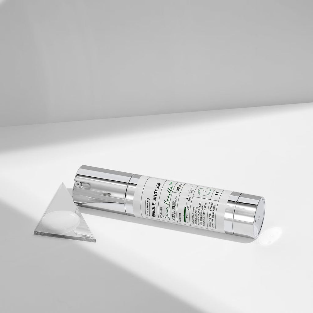 VT Reedle Shot 300 lies on a white surface with a triangular clear lid or insert beside it. The bottle has a white printed label with black text and small green accents, photographed in a clean studio setting with strong directional light and soft shadows.