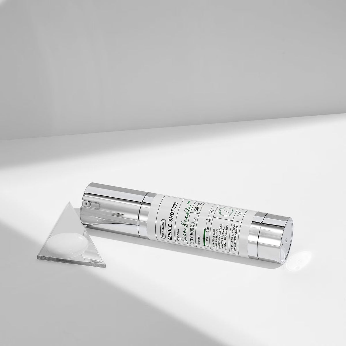 VT Reedle Shot 300 lies on a white surface with a triangular clear lid or insert beside it. The bottle has a white printed label with black text and small green accents, photographed in a clean studio setting with strong directional light and soft shadows.