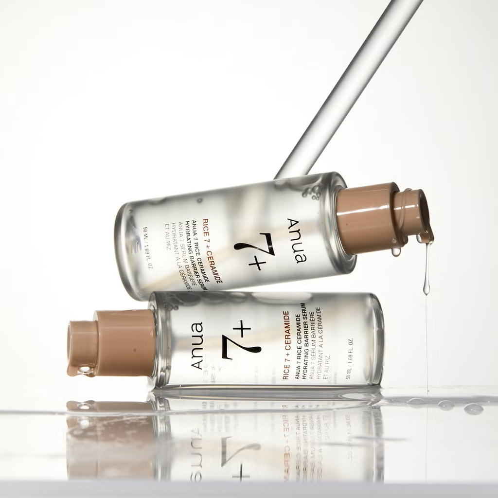 Two clear glass bottles of Anua 7 Rice Ceramide Hydrating Barrier Serum with beige pump dispensers, stacked and balanced on a glossy white reflective surface with a glass dropper above.