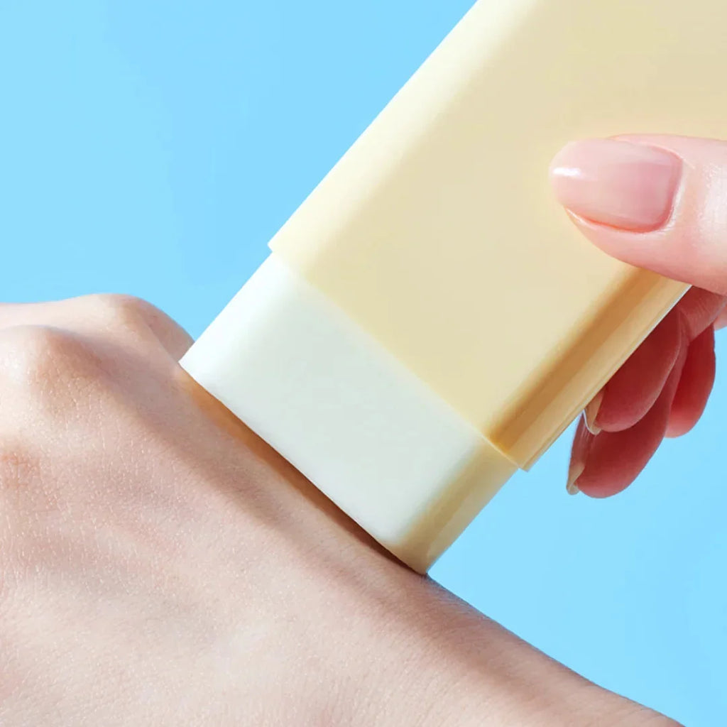 A close-up of a hand applying a pale Tocobo Vita Waterproof Sun Stick , onto the back of another hand against a solid light blue background.