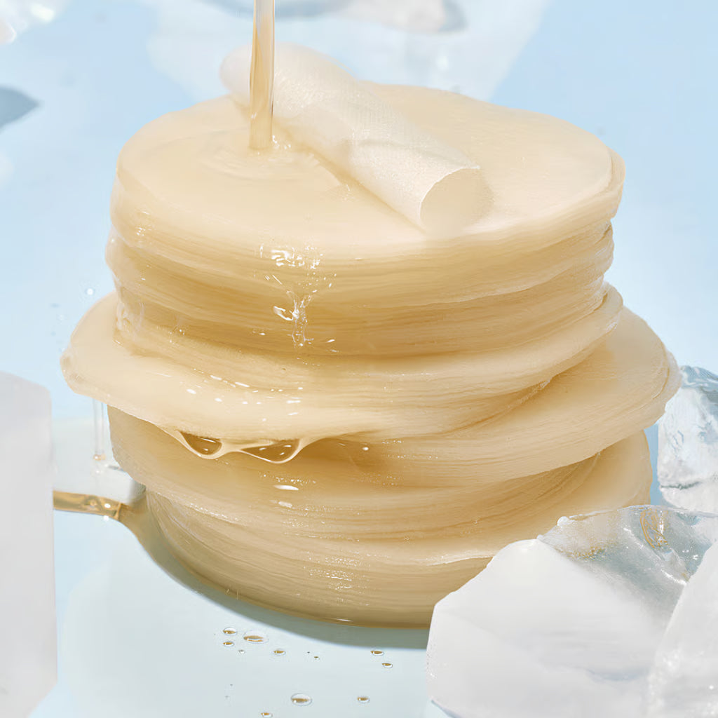 A stack of translucent, pale beige round Anua Hyaluronic Acid 8 + Catechin Cool Calming Pads, with a glossy liquid being poured over the top, set against a light blue background with ice cubes.